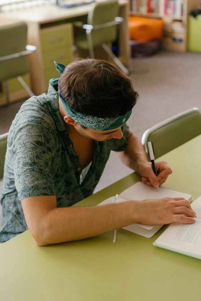 Man writing notes in a book at an indoor study area with bookshelves.